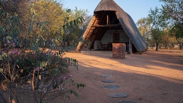 Un bâtiment à toit de chaume, doté de fondations en briques, entouré d’un chemin de terre et de végétation.
