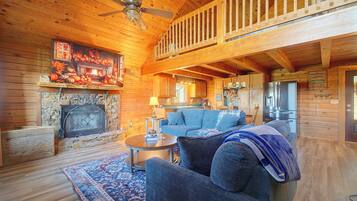 A cozy cabin interior with a fireplace, a dining area, and a living room furnished with a sofa and armchairs.