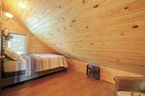 Ein Schlafzimmer mit schräg stehender Decke, Holzwänden, einem Bett mit gestreifter Decke, einer Nachttischlampe und einem Fenster mit Jalousien.