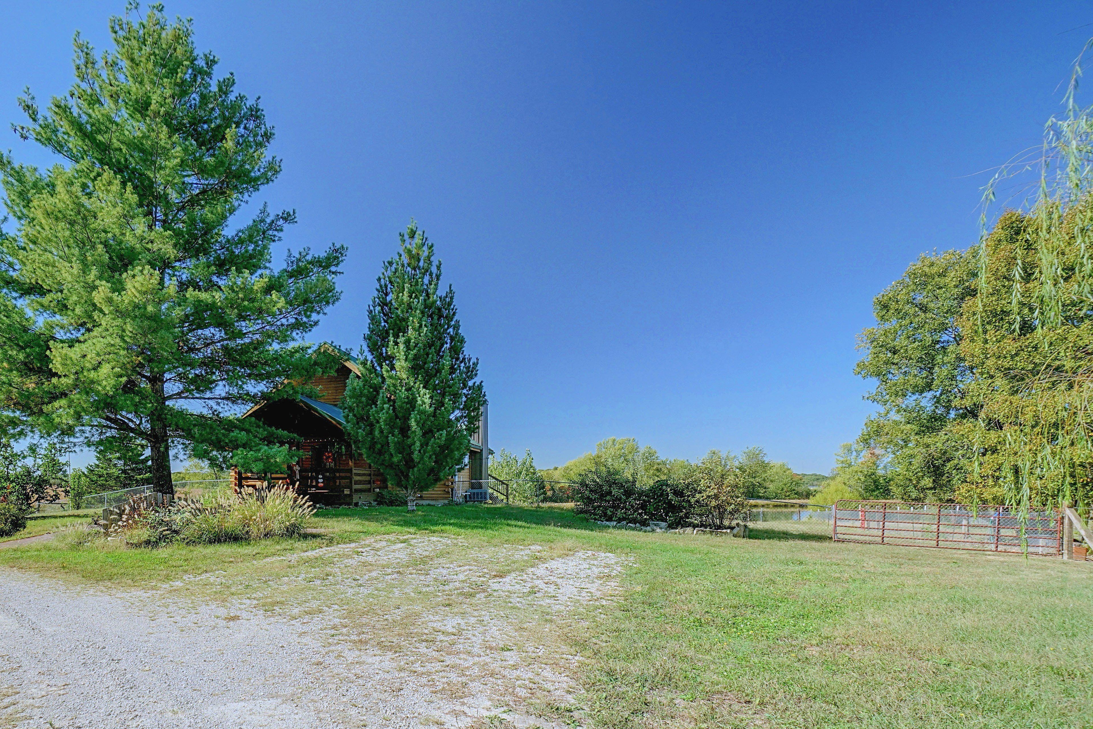 A rustic cabin with a gravel driveway, surrounded by trees and a fenced area.