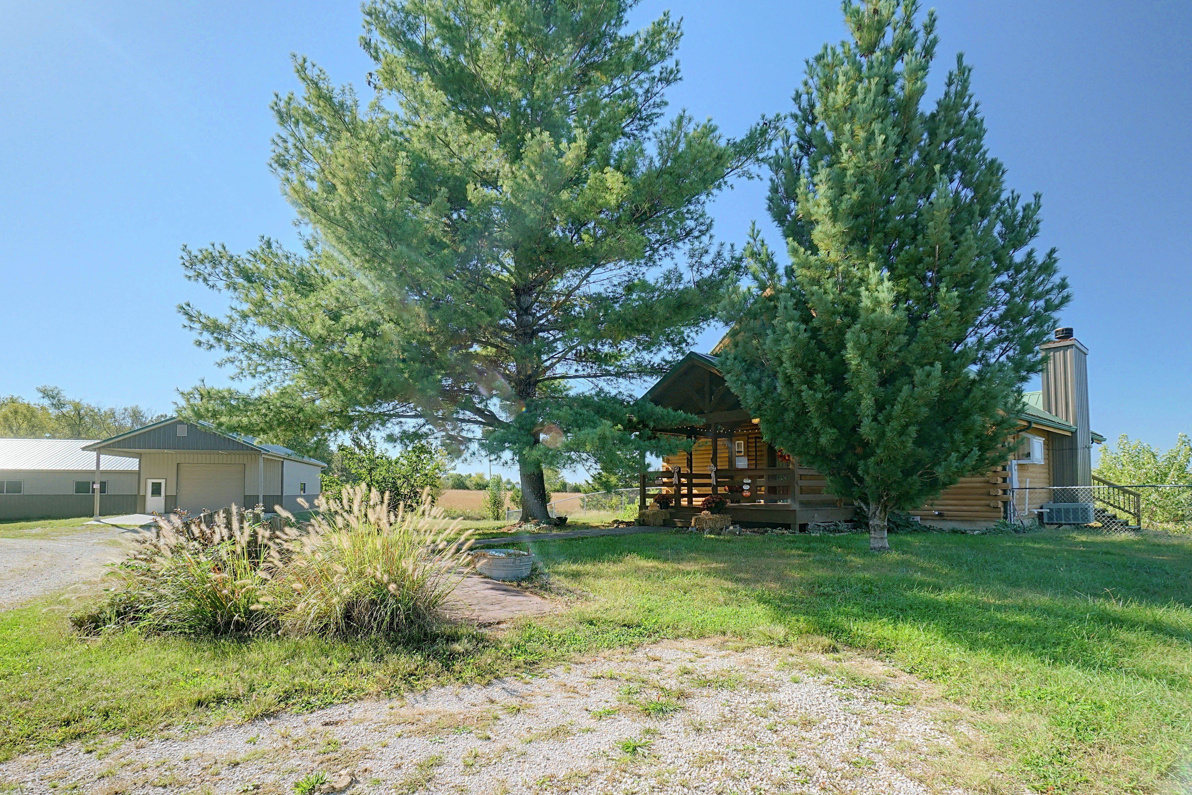 A cabin with a large tree in front, a gravel driveway, and a metal building in the background.