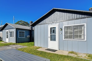 A small, single-story building with a front door and a large window, surrounded by a grassy area and a wooden deck. - Direct Shoreline Access! Tawas City Cabin (Tawas City)