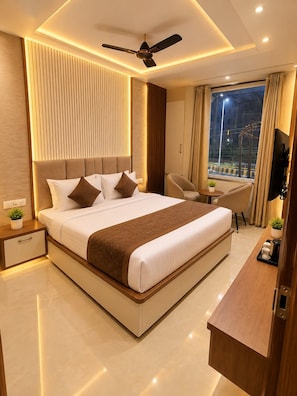 A modern hotel room with a large bed, a ceiling fan, a TV, and a view of the city at night. - Hotel Empire Inn  (Varanasi)