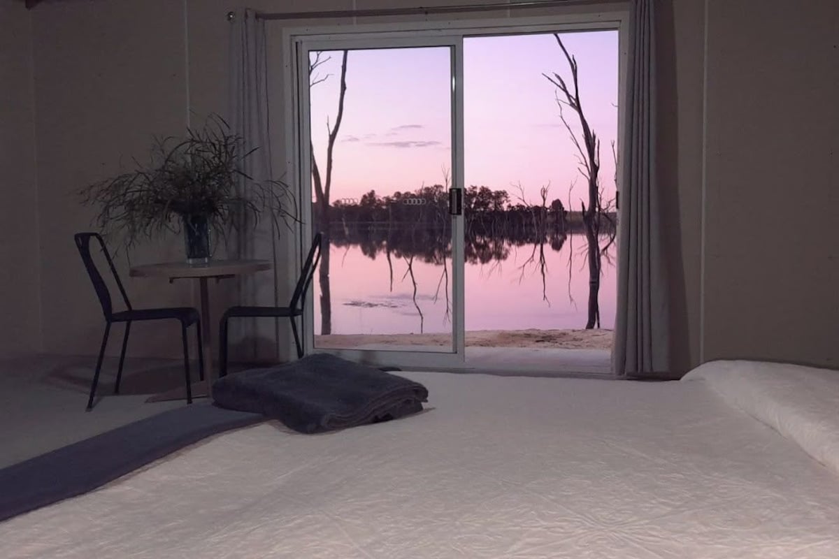 Una camera da letto con un letto grande, una sedia, un tavolino e vista su un lago tramite una porta scorrevole.