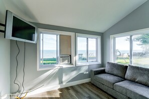 A room with a grey couch, a flat-screen TV mounted on the wall, and large windows offering a view of the outdoors. - Yard w/ Lake Views! Quaint Tawas City Escape (Tawas City)