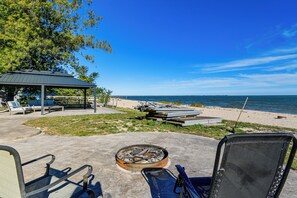 En terrasse ved stranden med et bålsted, liggestole og et overdækket siddeområde med udsigt over havet.