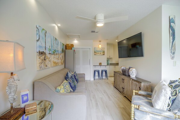 A modern living room with a sofa, a dining area with stools, and a kitchen in the background. - Cape Canaveral Getaway w/ Pool - Walk to Beach (Cape Canaveral)
