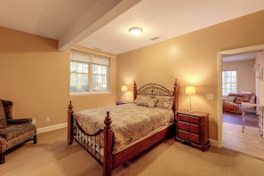 A bedroom with a wooden bed, two bedside lamps, a window with blinds, a chair, and a view of another room with a couch. - Quiet Horse Farm Hideaway Near Downtown Aiken (Aiken)