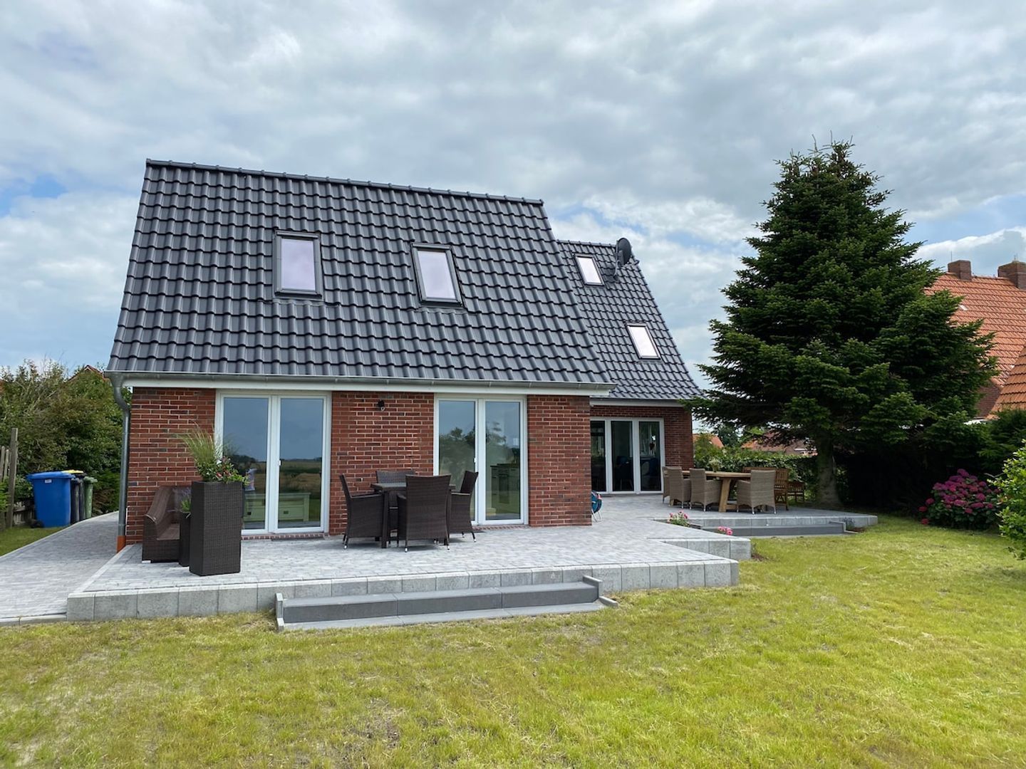 Ein zweistöckiges Haus mit geneigtem Dach, einer Terrasse mit Gartenmöbeln und einer Ziegelhausfassade.