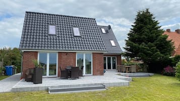 Ein zweistöckiges Haus mit geneigtem Dach, einer Terrasse mit Gartenmöbeln und einer Ziegelhausfassade.