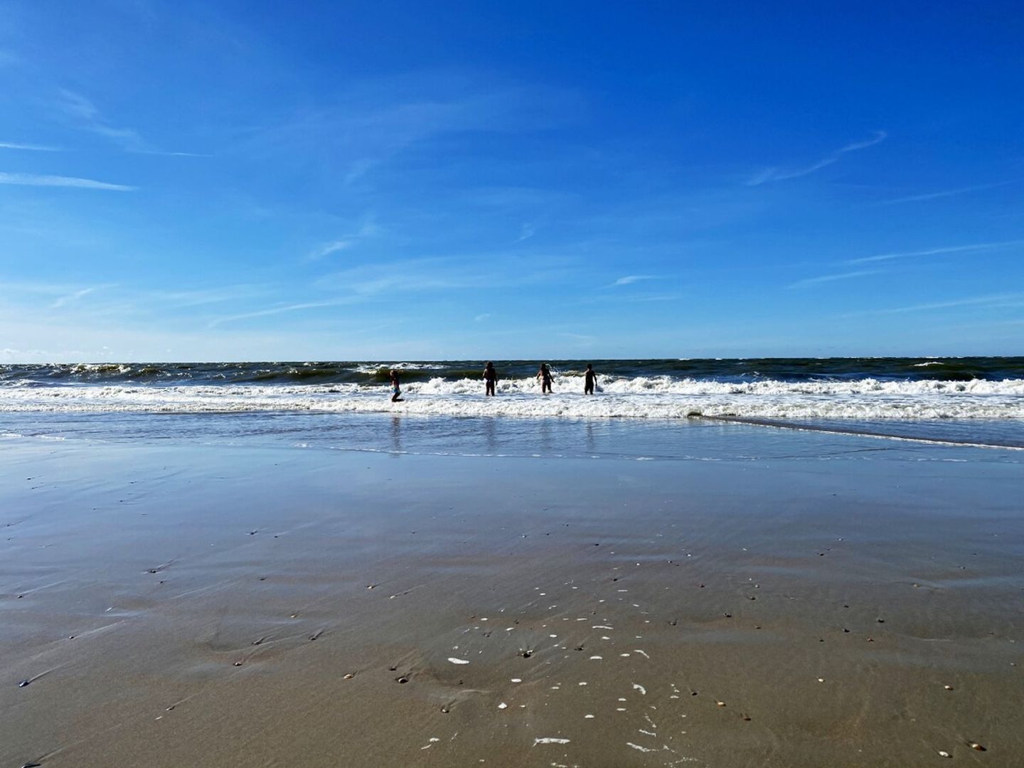 A beach with people walking along the shore, a clear sky, and waves approaching the beach.