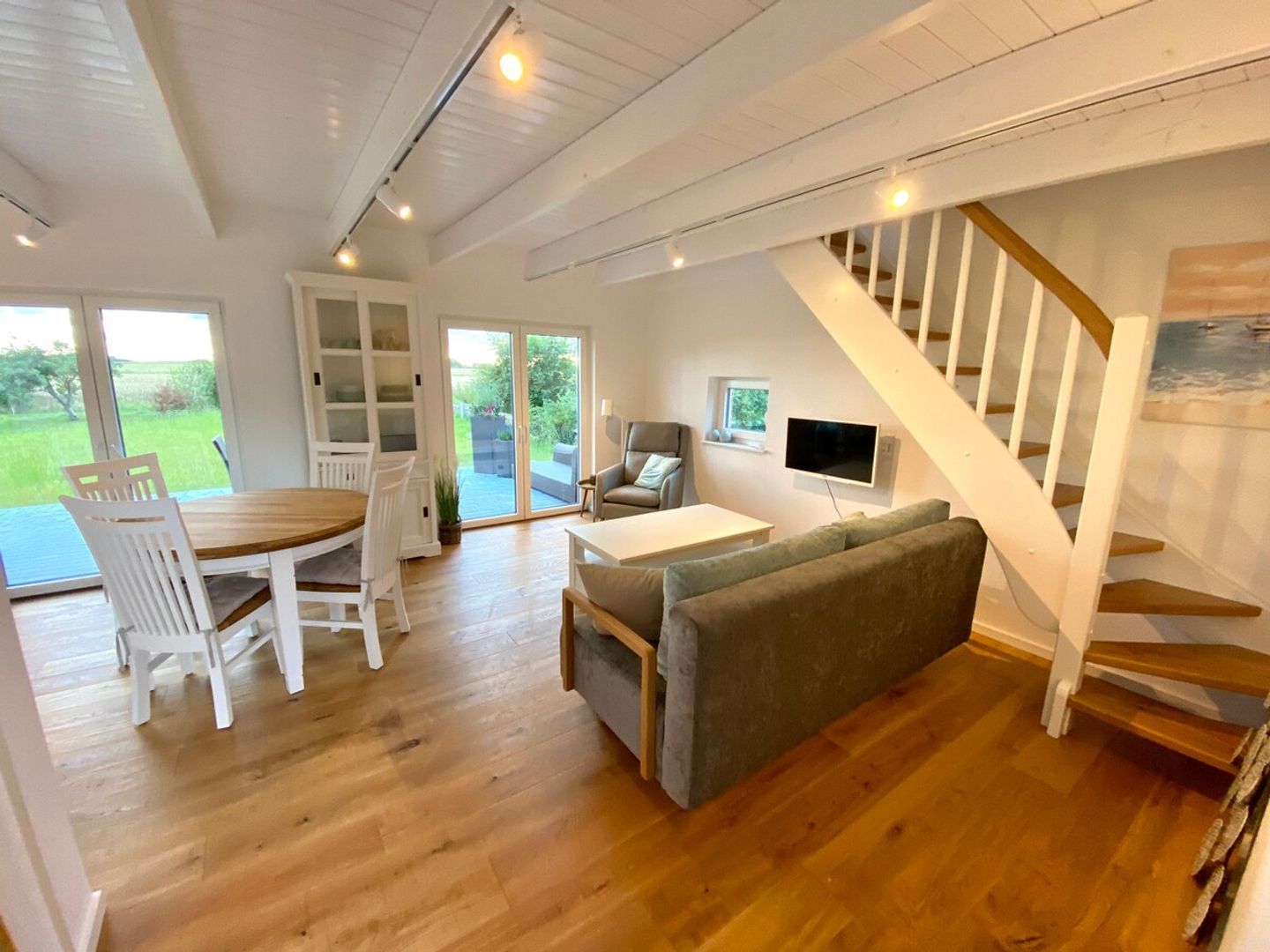A spacious living area with a wooden floor, a dining table with chairs, a sofa, a television, and a staircase.
