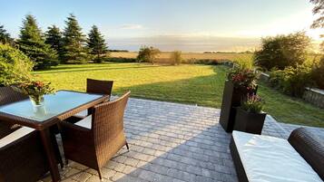 An outdoor patio with a glass-top table and wicker chairs, overlooking a grassy field and trees.