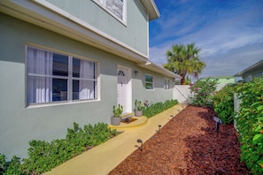 A building with a light green exterior, a window with white curtains, a door with a white frame, and a small garden area with plants and mulch. - Pool Access, Walk to Beach! Cape Canaveral Apt (Cape Canaveral)