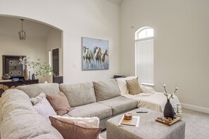 A living room with a large sofa, a coffee table, a painting of horses, a vase with flowers, and a window with blinds. - Yard, Patio, Office! Spacious Home in Union Park (Little Elm)