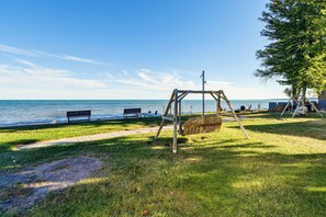 Un parc avec une aire de jeux comprenant des balançoires, des bancs, un sentier et une étendue d’eau en arrière-plan.