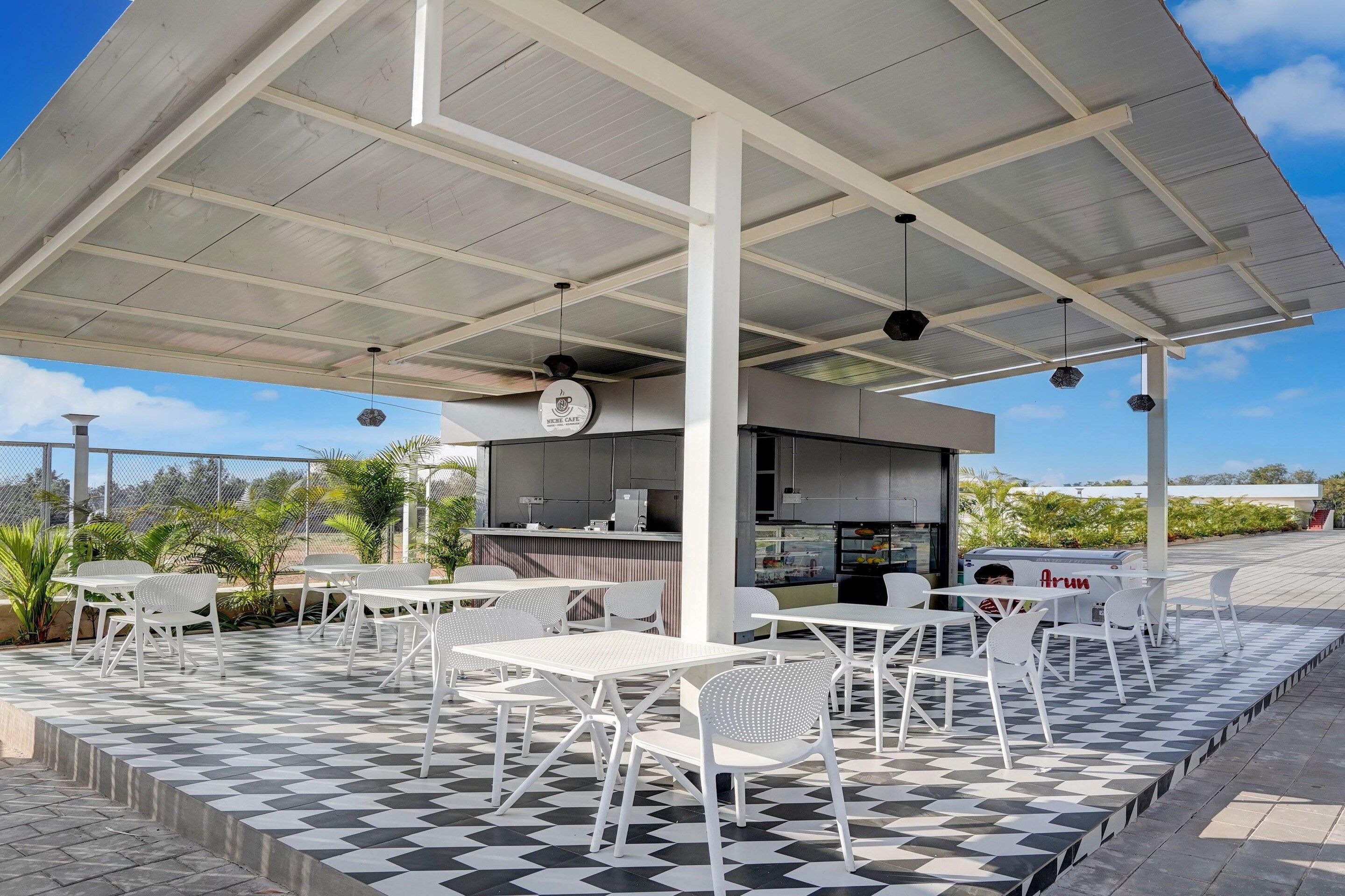 An outdoor cafe with a checkered floor, white tables and chairs, and a covered seating area.