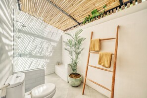 Une salle de bain avec des toilettes, un lavabo et une douche. On y trouve une échelle en bois sur laquelle sont posées des serviettes, ainsi qu’une plante en pot.