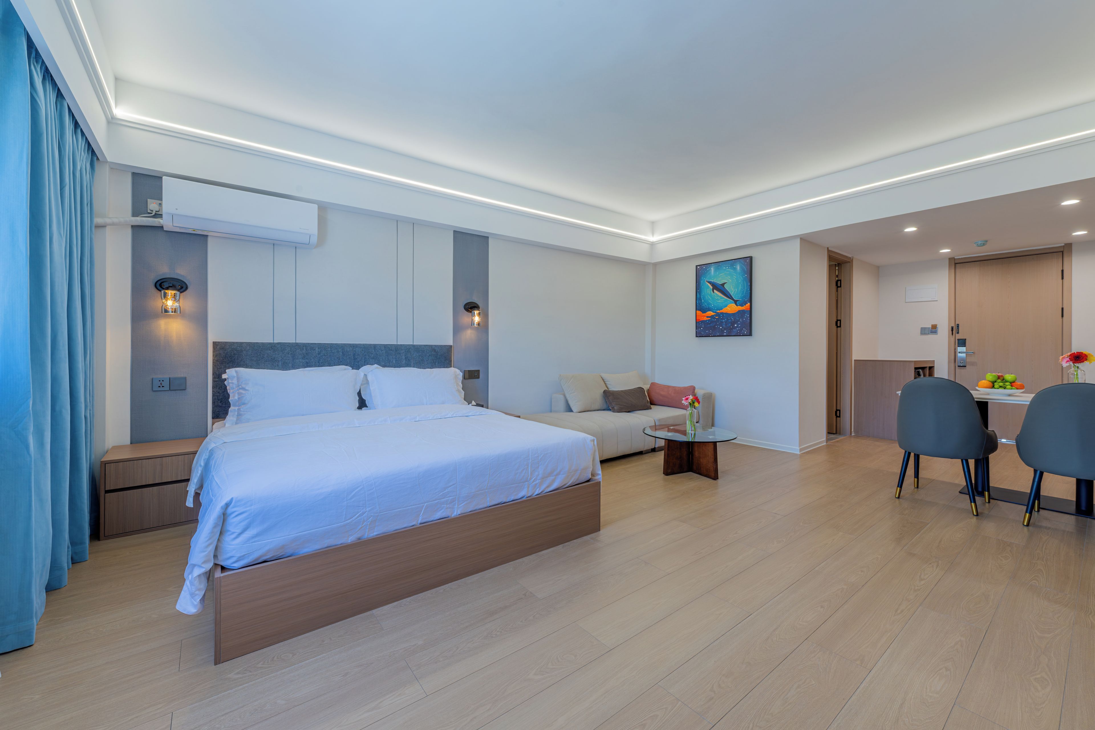 A modern hotel room with a large bed, a sofa, a dining table with chairs, and a painting on the wall.