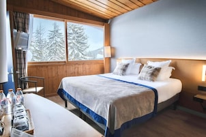 A hotel room with a bed, a table with water bottles, a chair, and a window with a view of snow-covered trees.
