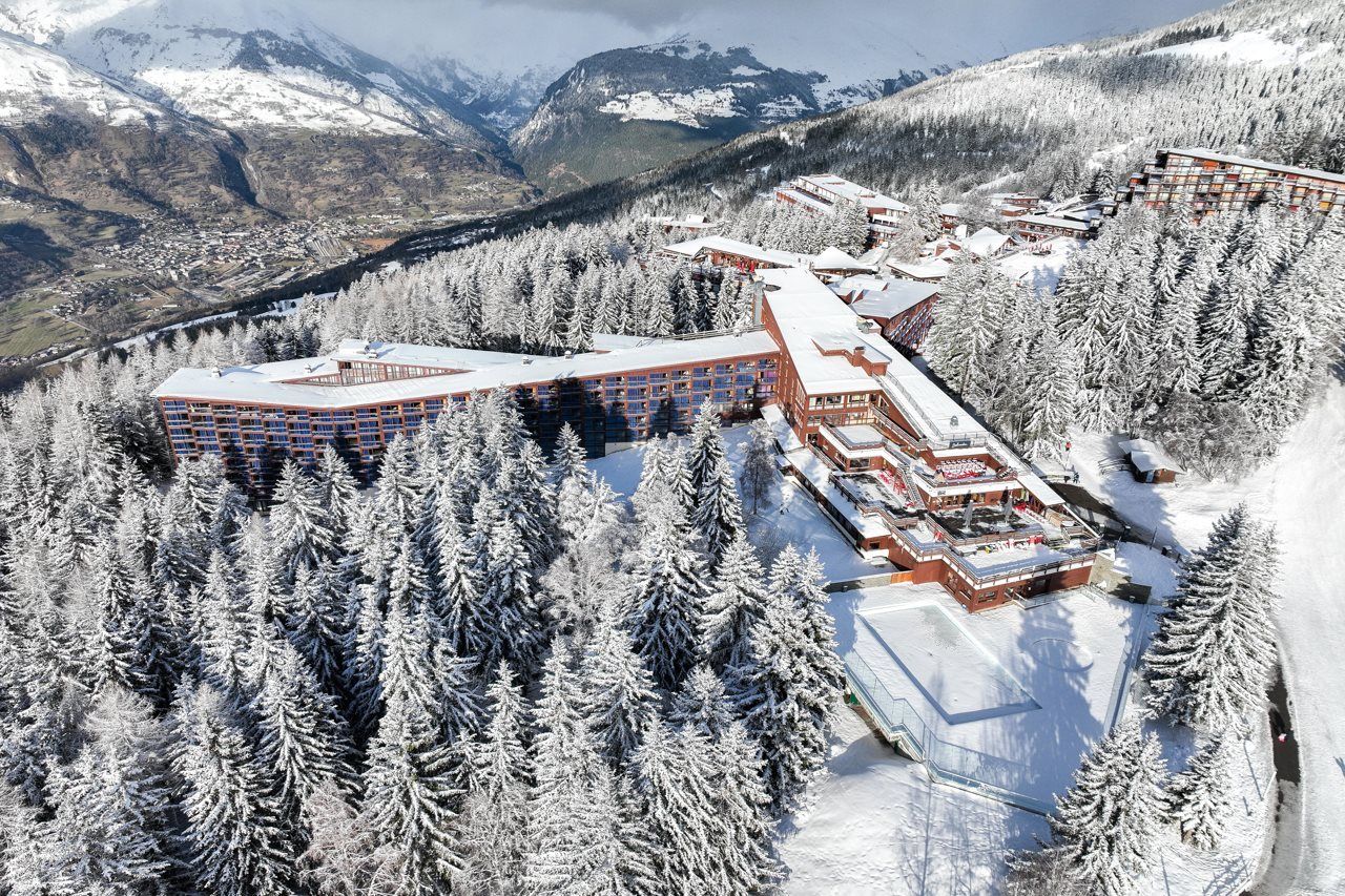 A snow-covered resort with multiple buildings, surrounded by dense forests and mountains in the background.