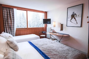 Ein Hotelzimmer mit zwei Betten, einem Schreibtisch mit Stuhl, einem Fernseher und einem Fenster mit Vorhängen.