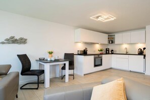 A modern kitchen with white cabinetry, a dining area with a table and chairs, and a living room with a sofa. - Holiday apartment with 1 bedroom (Westerland)