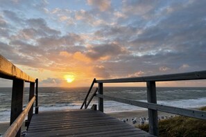 Ein Holzsteg führt zu einem Strand, im Hintergrund geht die Sonne unter.