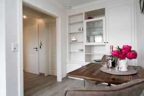 A dining area with a wooden table, chairs, a white cabinet with glass doors, and a vase with flowers. - Holiday apartment with 1 bedroom (Westerland)