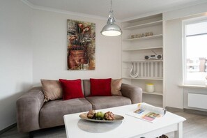 A living room with a grey sofa, red cushions, a white coffee table with a bowl of fruit, a painting on the wall, and a white shelving unit with a radio. - Holiday apartment with 1 bedroom (Westerland)