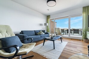 A modern living room with a sofa, armchair, coffee table, and a view of the outdoors through large sliding doors. - Apartment in Olpenitz with swimming pool (Olpenitz)