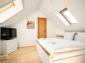 Quarto com teto inclinado, cama com roupa de cama branca, televisão sobre um móvel branco e porta de madeira.