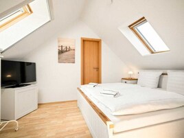 Un dormitorio con techo inclinado, una cama con ropa blanca, un televisor sobre un mueble blanco y una puerta de madera.