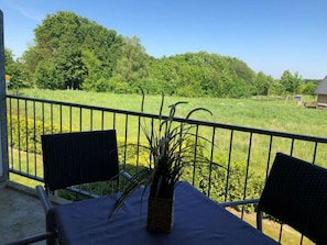 Varanda com duas cadeiras e uma mesa, com vista para uma área gramada com árvores e um pequeno edifício.