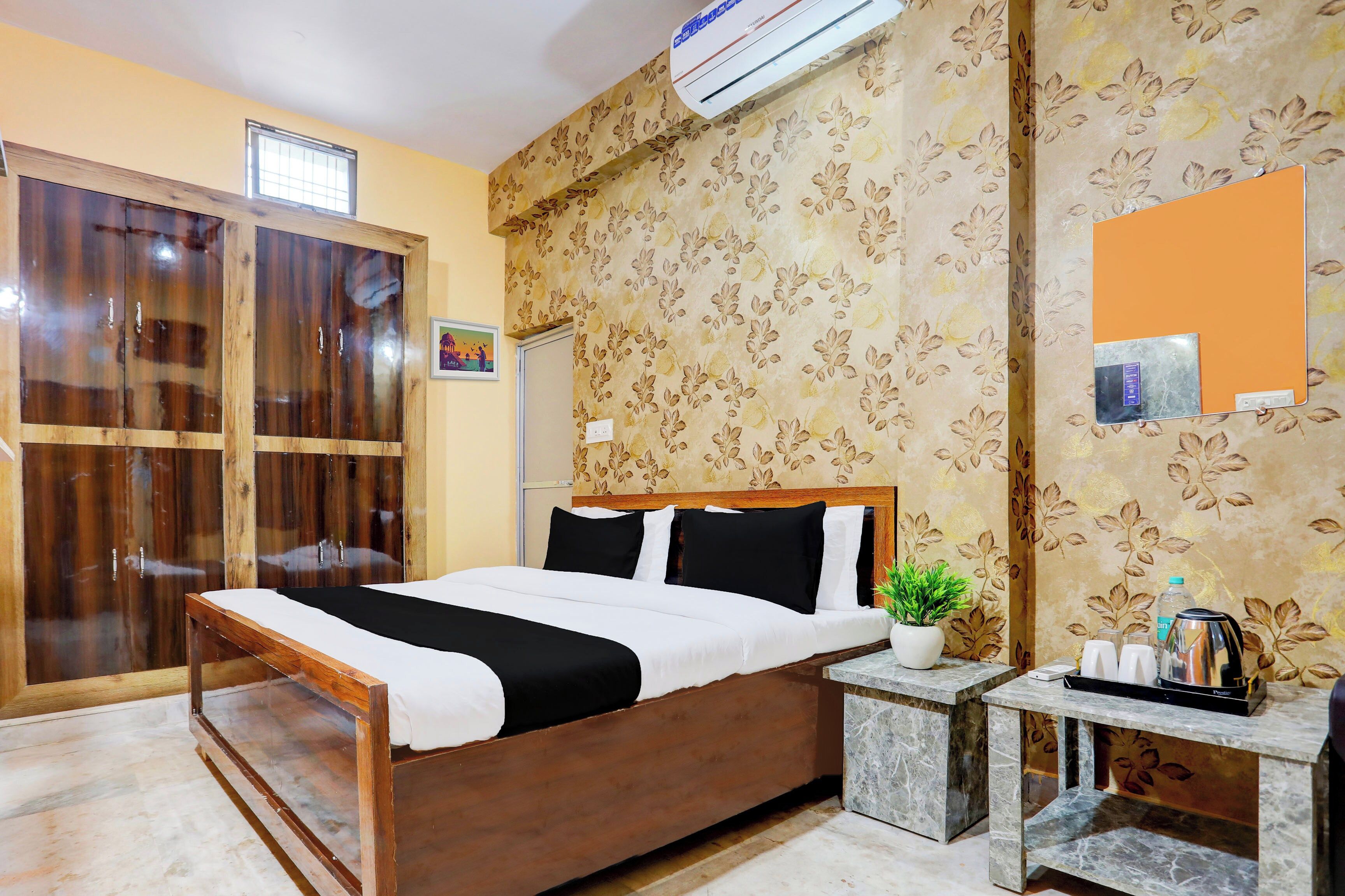 A hotel room with a wooden bed, a small bedside table, a potted plant, and a wall-mounted air conditioner.