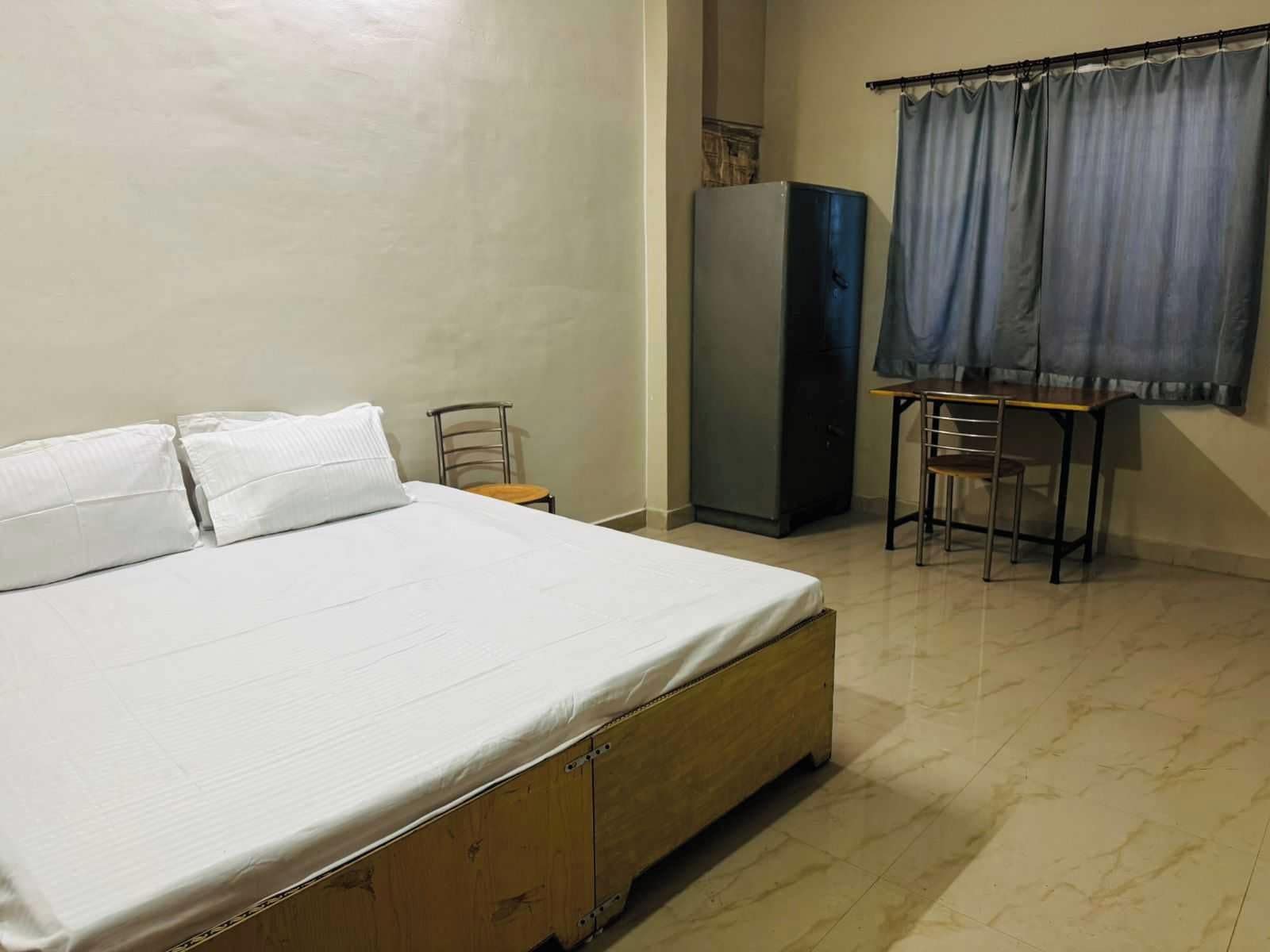 Une chambre à coucher avec un lit, une petite table, une chaise, une armoire et une fenêtre avec des rideaux.