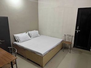 Une petite chambre avec un lit simple, un cadre de lit en bois, une chaise en métal et une table en bois.