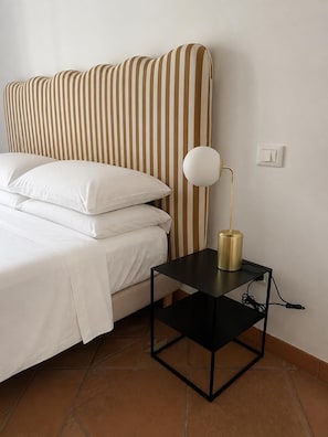 A bed with white bedding and a striped headboard, a black nightstand with a gold lamp, and a power outlet on the wall. - New Listing (Roma)
