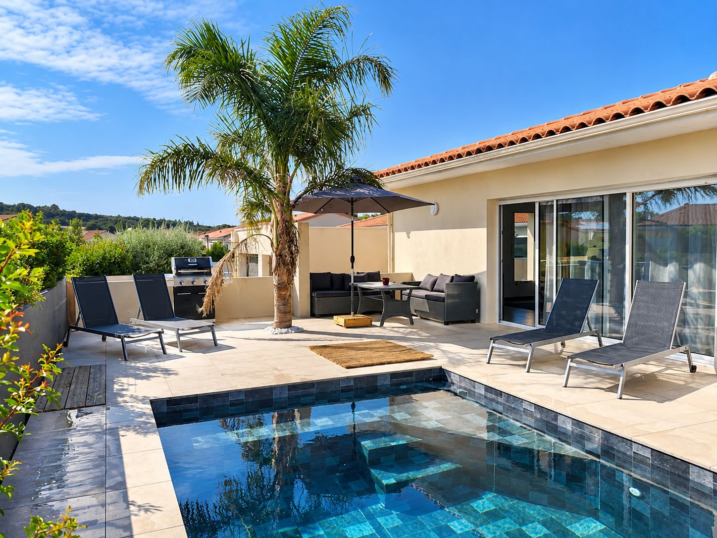 Une piscine avec un bord carrelé, un palmier, du mobilier d’extérieur et une porte-fenêtre coulissante.