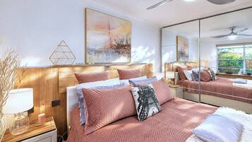 A bedroom with a wooden headboard, a bed with pink bedding, a bedside table with a lamp, and a large mirror reflecting the room.