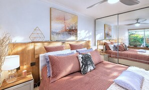 A bedroom with a wooden headboard, a bed with pink bedding, a bedside table with a lamp, and a large mirror reflecting the room.