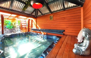 A hot tub with a wooden bench and a Buddha statue, surrounded by wooden walls and a metal roof.