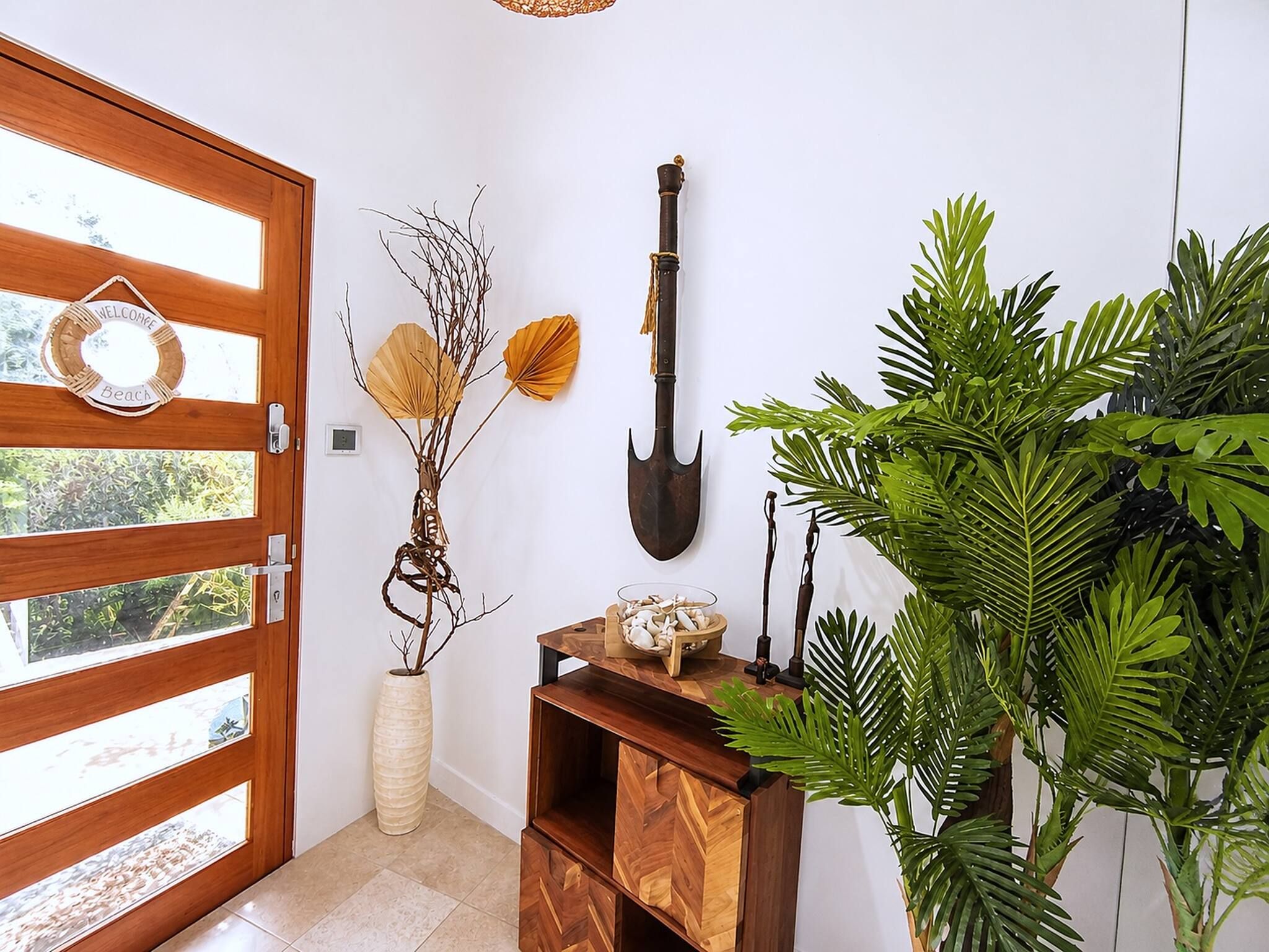 Une porte en bois ornée d’une pancarte « Bienvenue », une pelle fixée au mur, un vase rempli de branches et un meuble en bois contenant un bol rempli de coquillages.
