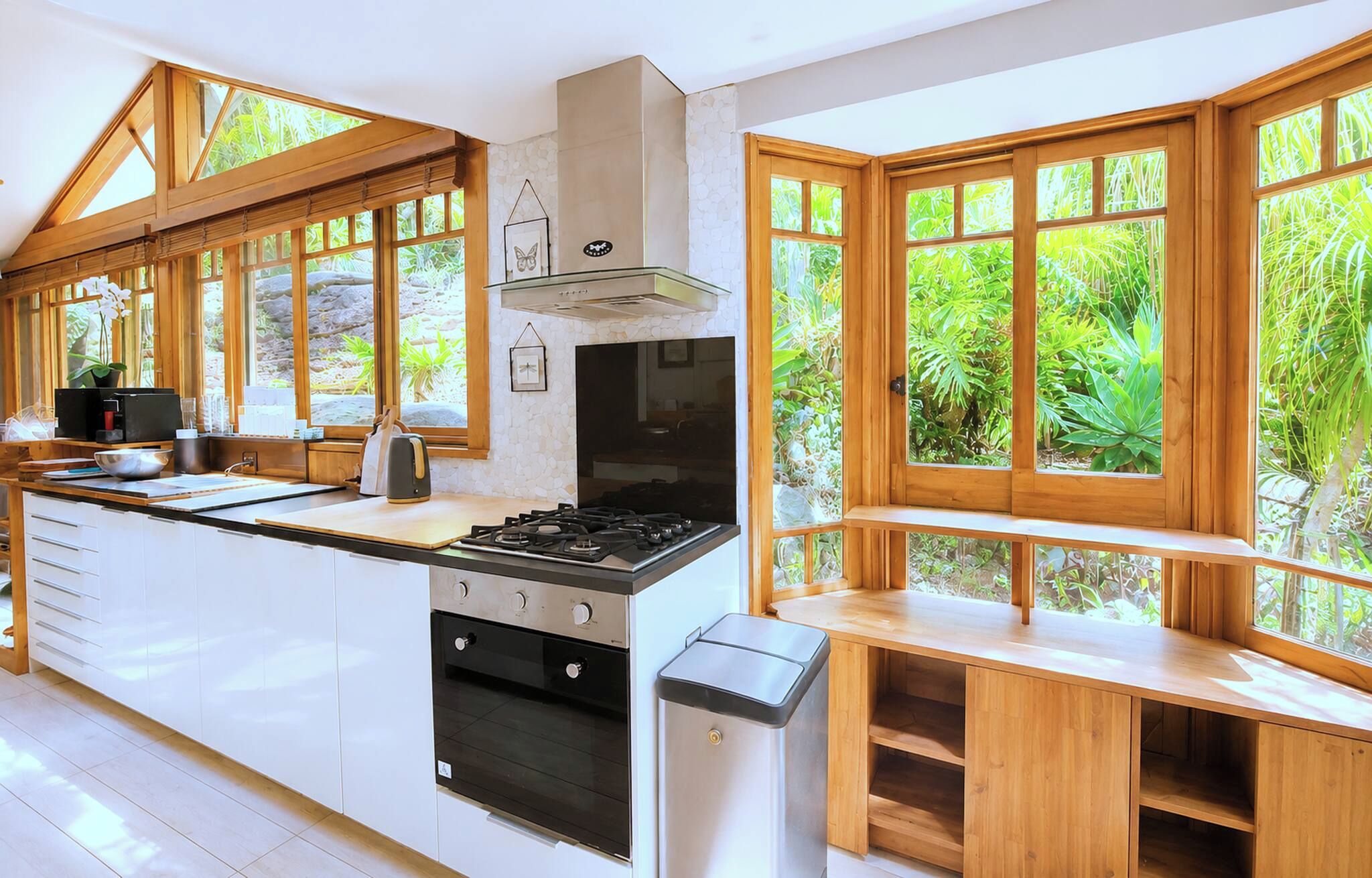 Une cuisine dotée d’un grand îlot central, de meubles en bois, d’une cuisinière à gaz et offrant une vue sur la verdure grâce à de grandes fenêtres.