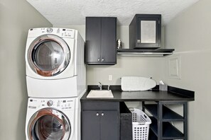 A laundry room with two stacked front-loading washing machines, a sink, a drying rack, and a small cabinet. - Eight Seventeen Abode | Near UM & Downtown (Missoula)