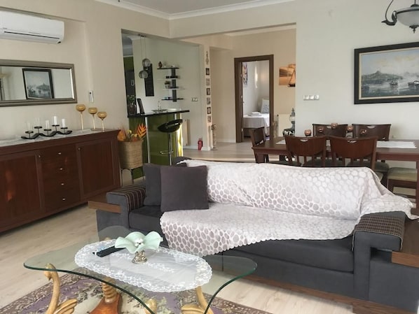 Ein Wohnzimmer mit einer Couch, einem Glastisch, einem Essbereich mit Stühlen und einer Küche im Hintergrund.