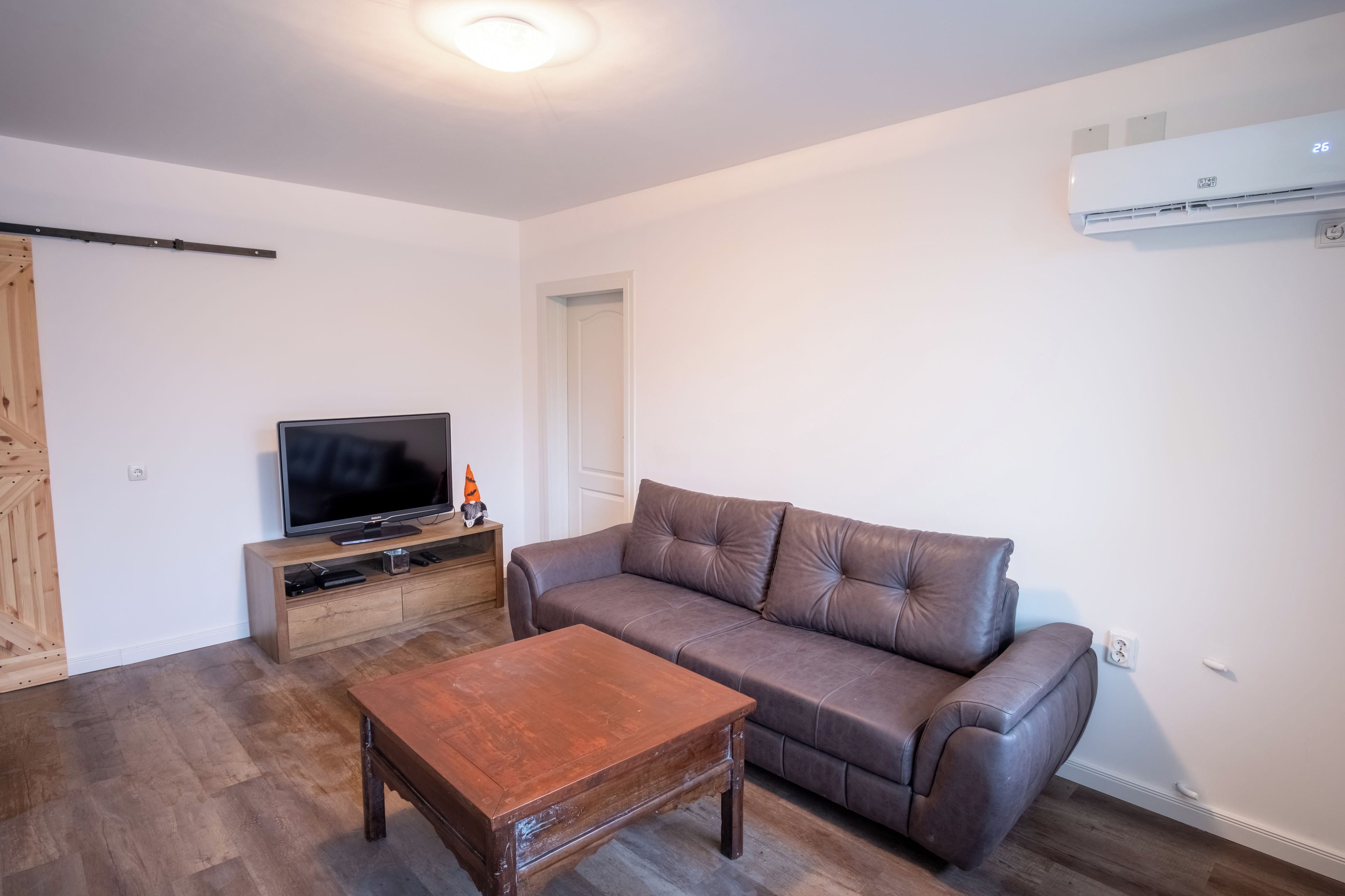 A living room with a brown leather sofa, a wooden coffee table, a TV on a wooden stand, and an air conditioning unit on the wall.