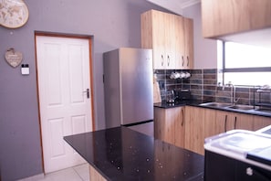 A kitchen with a refrigerator, wooden cabinets, a black countertop, and a window. - Langa Elihle on Paradise Beach (Jeffreys Bay)