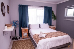 A bedroom with a bed, a bedside table, a fan, a small cabinet, and a window with curtains. - Langa Elihle on Paradise Beach (Jeffreys Bay)