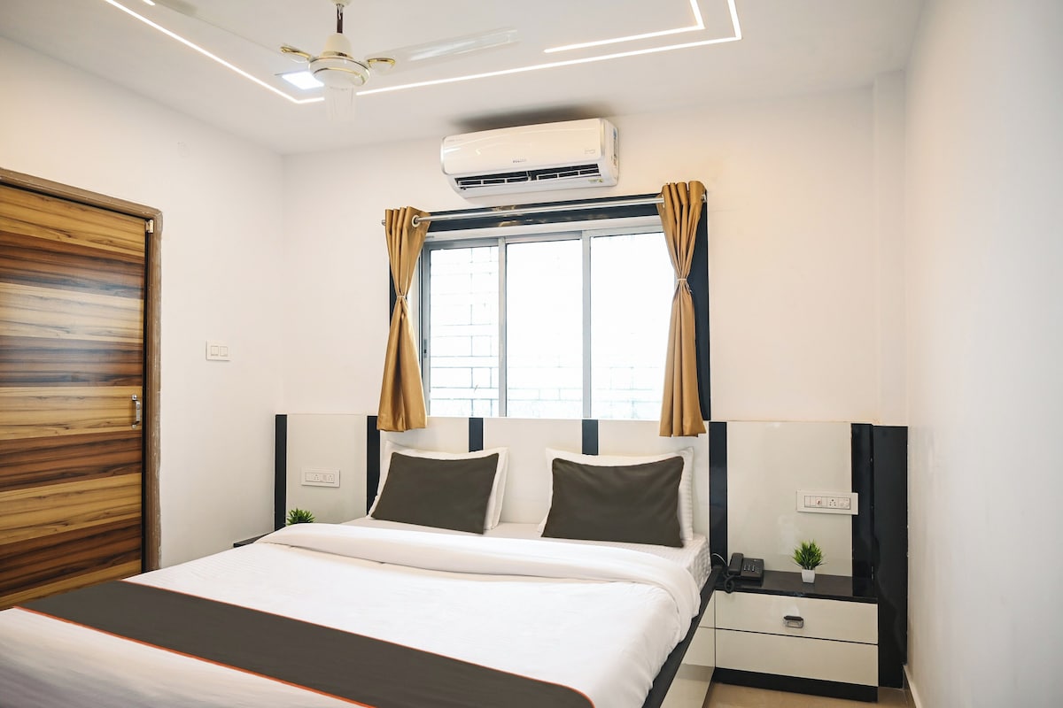 A hotel room with a bed, bedside tables, an air conditioning unit, a window with curtains, and a ceiling light fixture.